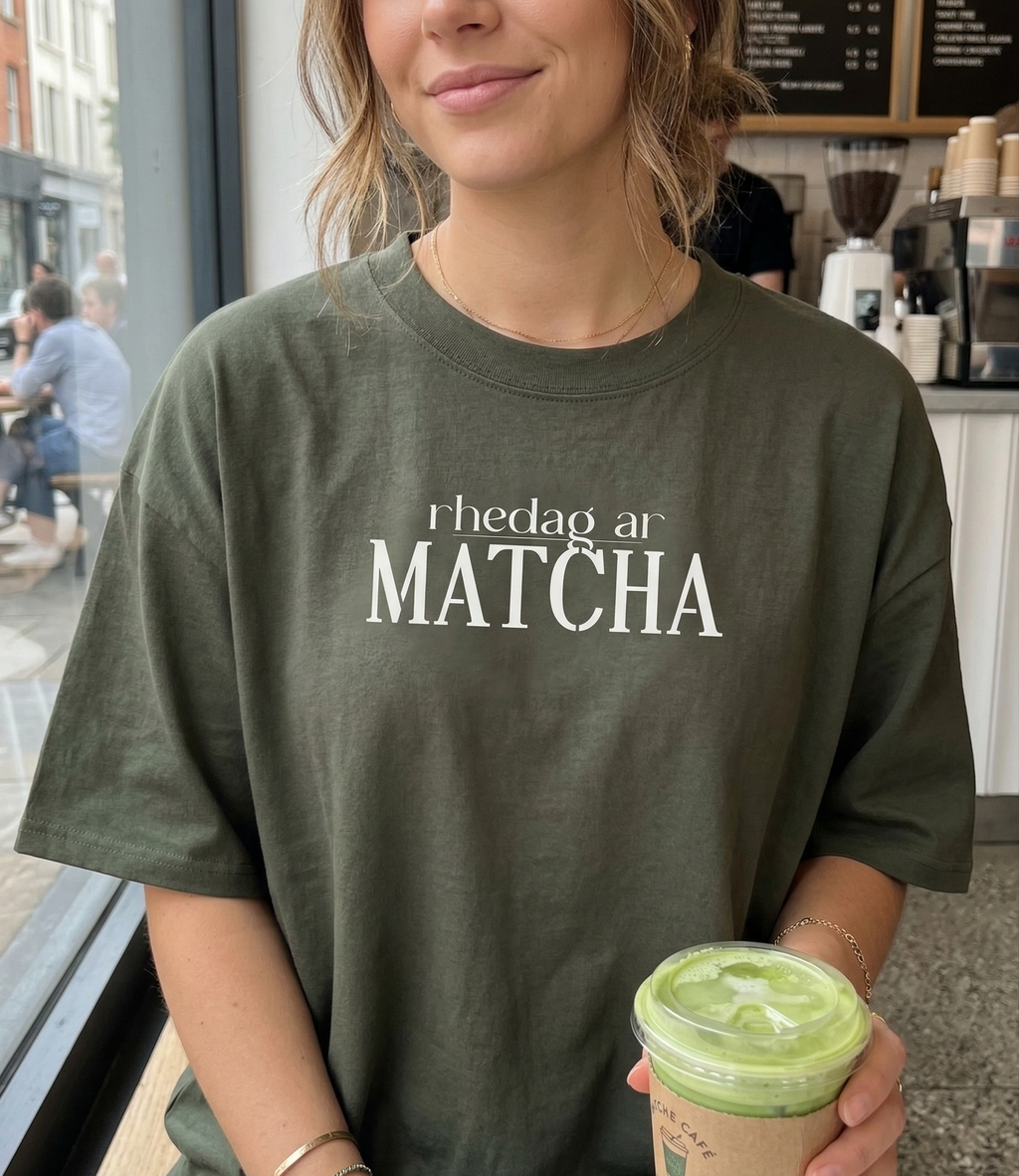 Person wearing a green t-shirt with 'rhedag ar Matcha' text, holding a matcha drink in a cafe.