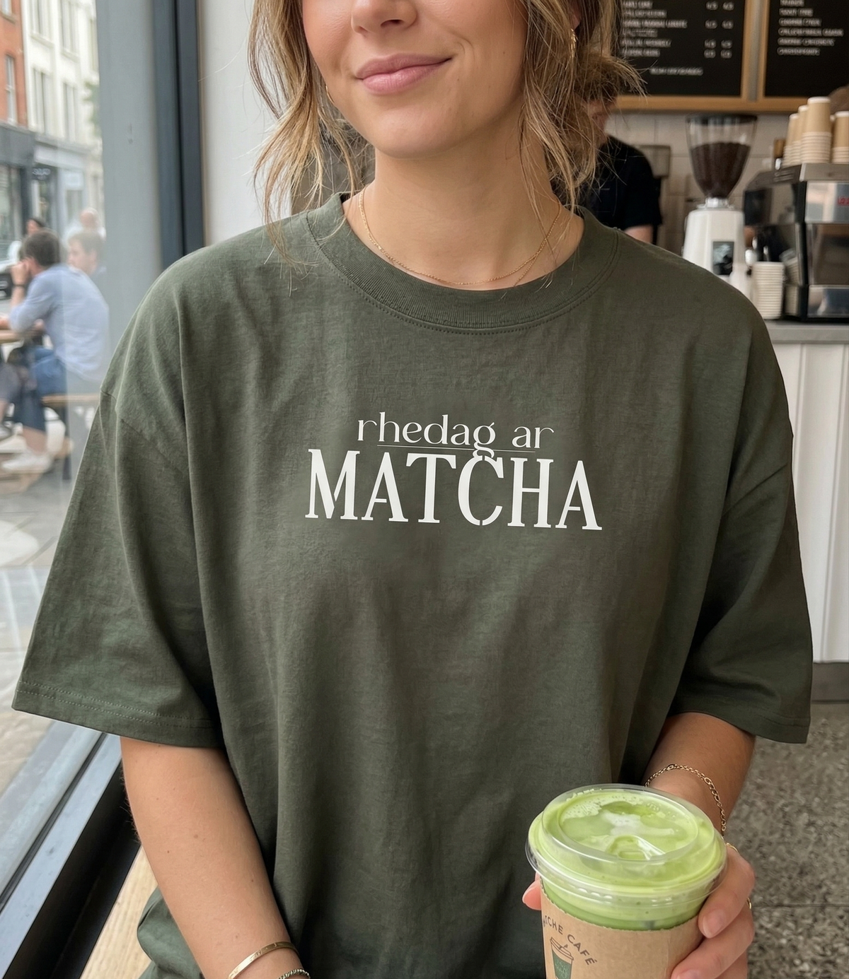Person wearing a green t-shirt with 'rhedag ar Matcha' text, holding a matcha drink in a cafe.